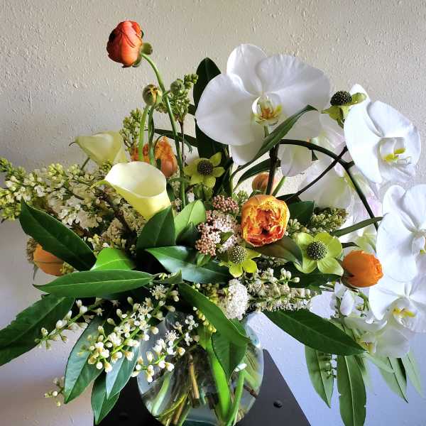 Arrangement of white orchids, yellow calla lilies, and orange ranunculus in a clear glass vase