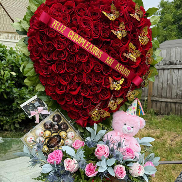 Heart-shaped red rose arrangement with pink roses, teddy bear, and chocolates
