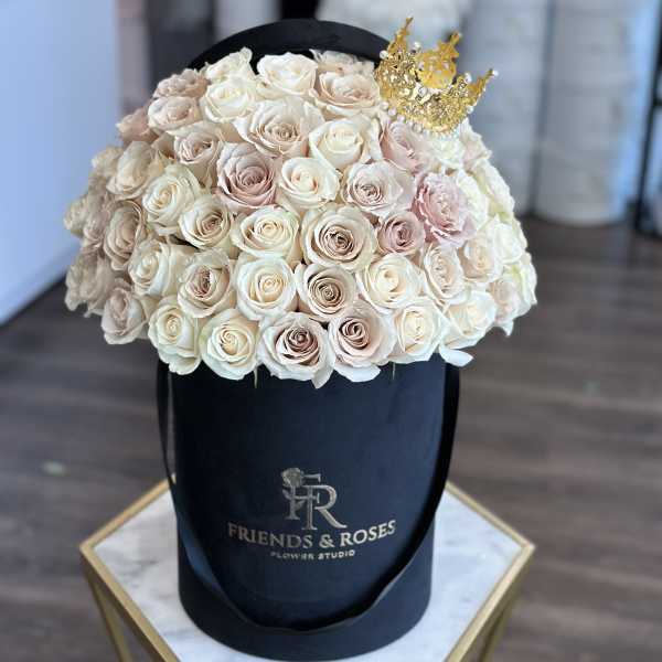 Cream and blush roses arranged in a black hat box with a gold crown topper.