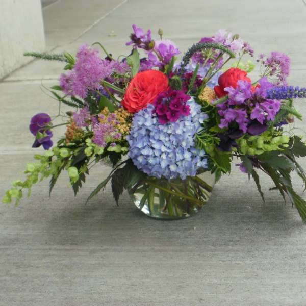 Low glass vase of mixed purple, blue, and red flowers arranged in a wide garden-style centerpiece