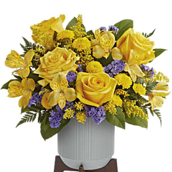 Yellow roses and yellow blooms with purple accents in a white ribbed pot on a wooden stand