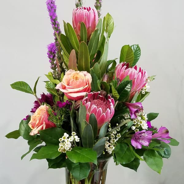 Tall arrangement of pink protea, roses, and purple flowers in a clear glass vase
