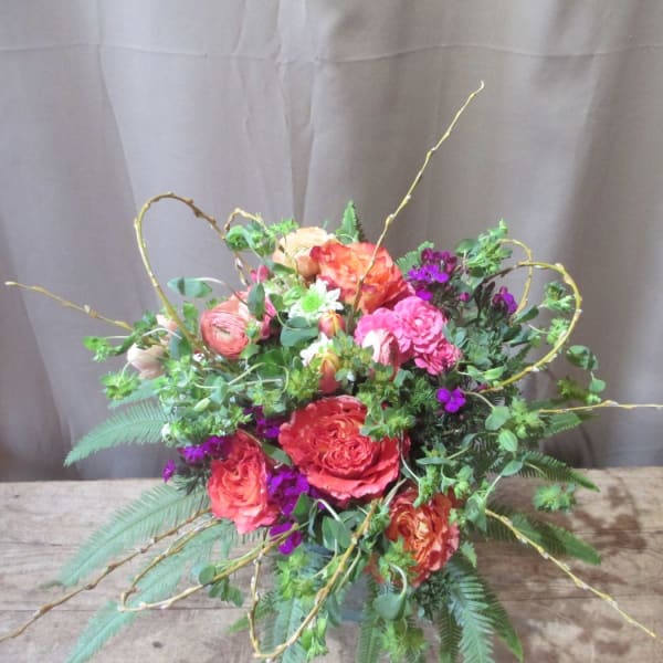 Colorful arrangement of orange and pink roses with purple flowers and curly branches in a low vase