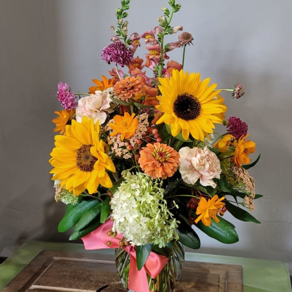 Tall mixed arrangement with sunflowers, hydrangea, and assorted orange and peach blooms in a glass vase.