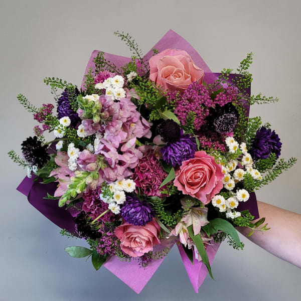 Hand-tied bouquet of pink roses, purple flowers, and white daisies wrapped in pink and purple paper