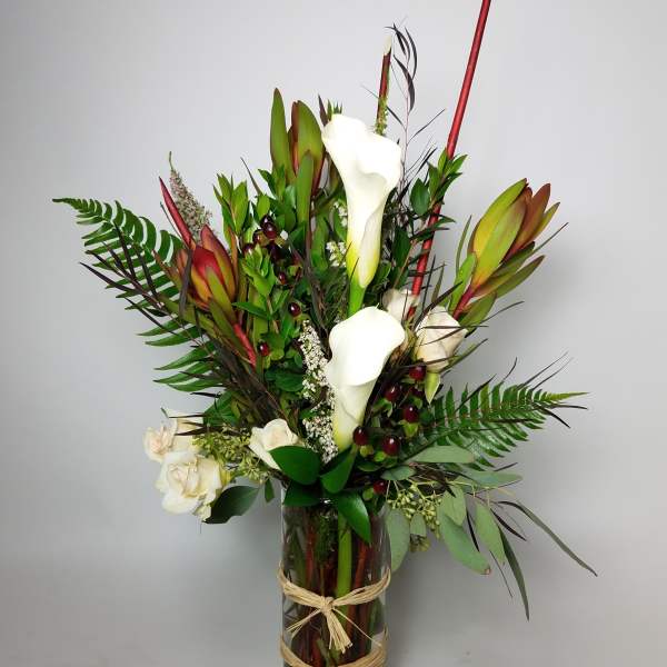 Tall arrangement of white calla lilies and red tropical blooms in a glass vase