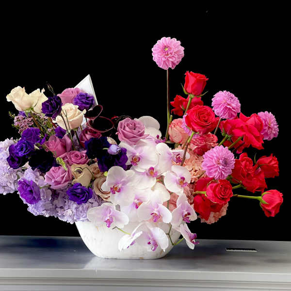 Asymmetrical bowl arrangement of pink, purple and red roses, dahlias and orchids in a white vessel.