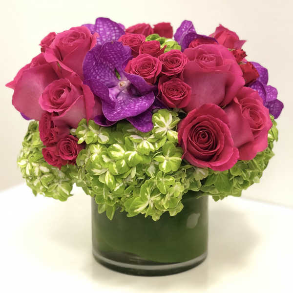 Low arrangement of hot pink roses, purple orchids, and green hydrangeas in a clear glass cylinder vase