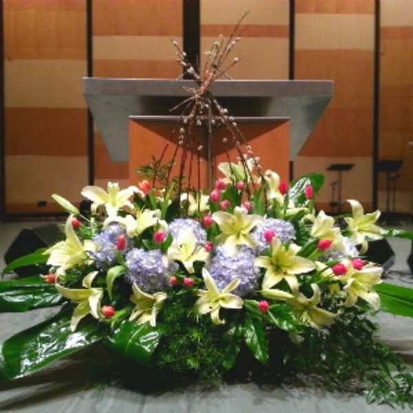 Low altar arrangement of yellow lilies, lavender hydrangeas, and pink buds with large glossy foliage.