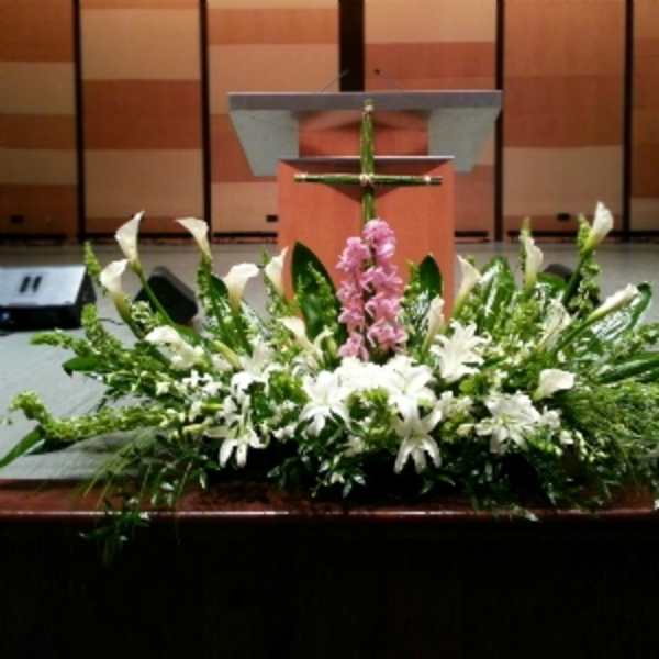 Large white and green altar arrangement with lilies, calla lilies, and a pink flower cluster beneath a green cross.