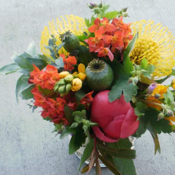 Bright mixed bouquet with yellow pincushion protea, orange blooms, and a pink peony in a glass vase