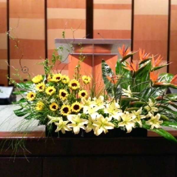 Long stage arrangement of yellow lilies, sunflowers, and orange tropical flowers in a low container