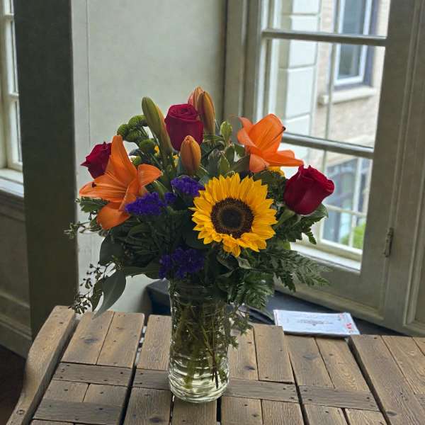 Bouquet of sunflowers, red roses, orange lilies, and purple filler in a glass vase