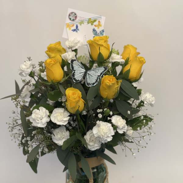 Yellow roses and white carnations in a glass vase with a butterfly pick