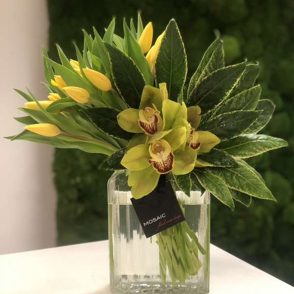 Yellow tulips and green orchids arranged with lush foliage in a clear glass vase