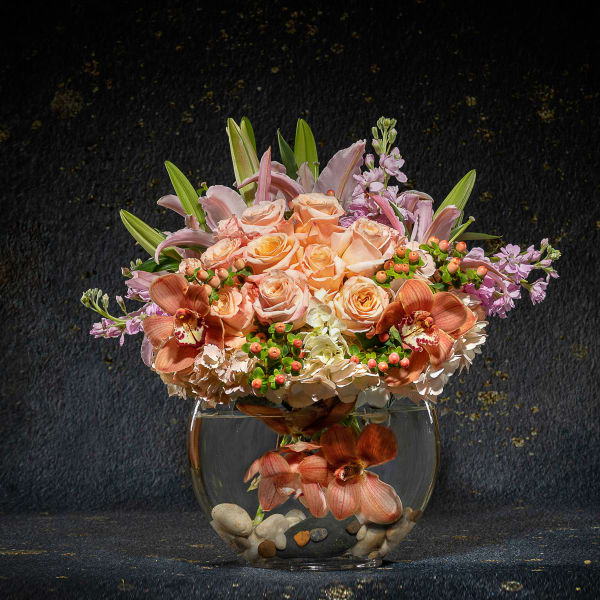 Peach roses and orchids arranged in a round glass vase with stones
