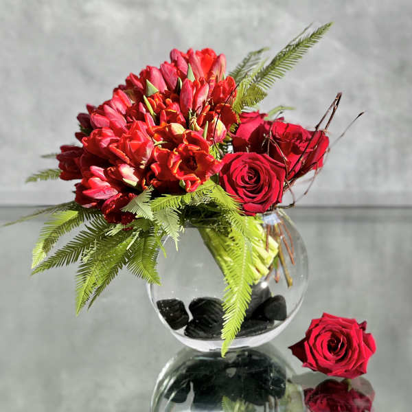 Red roses and tulips arranged in a round glass vase with fern foliage.