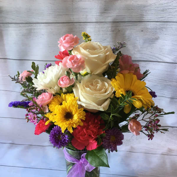 Mixed bouquet of roses, daisies, and carnations in a glass vase with a purple ribbon