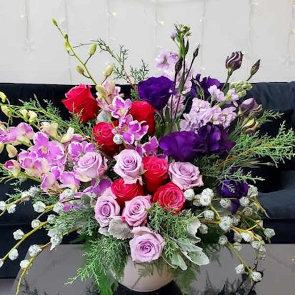Low bowl arrangement of pink and red roses with purple blooms and orchids in a lush mixed design