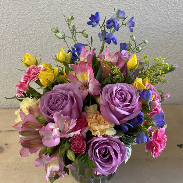 Mixed bouquet of purple roses, pink alstroemeria, and yellow blooms in a glass vase