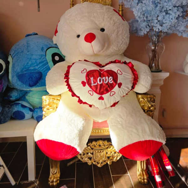 Large teddy bear holding a heart pillow beside a vase of blue flowers