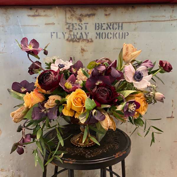 Mixed bouquet of purple, peach, and yellow flowers in a gold vase