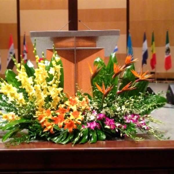 Large tropical stage arrangement with yellow spikes, orange lilies, and birds of paradise before a podium