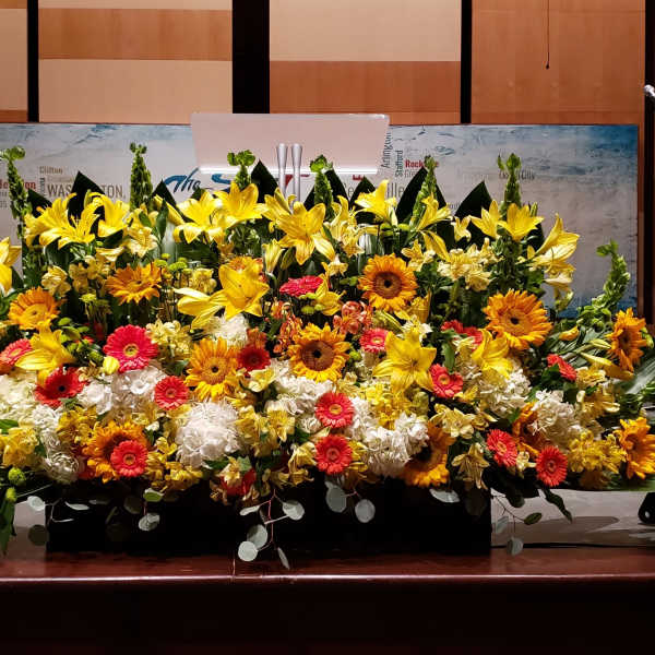 Large stage arrangement of yellow lilies, orange gerbera daisies, and white hydrangeas in a low black container