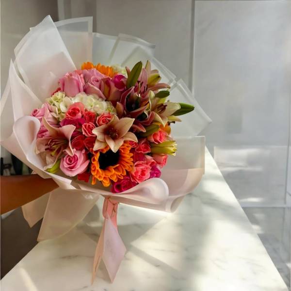 Hand-tied bouquet of pink roses, lilies, and orange flowers wrapped in white paper