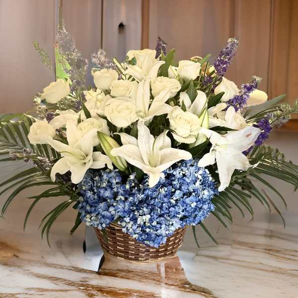 White lilies and roses arranged with blue hydrangeas in a wicker basket
