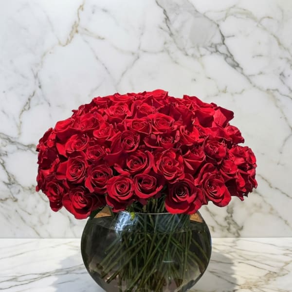 Round arrangement of red roses in a clear glass bowl vase with scattered rose petals.
