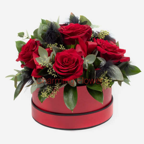 Red roses arranged in a round red box with greenery and black feathers