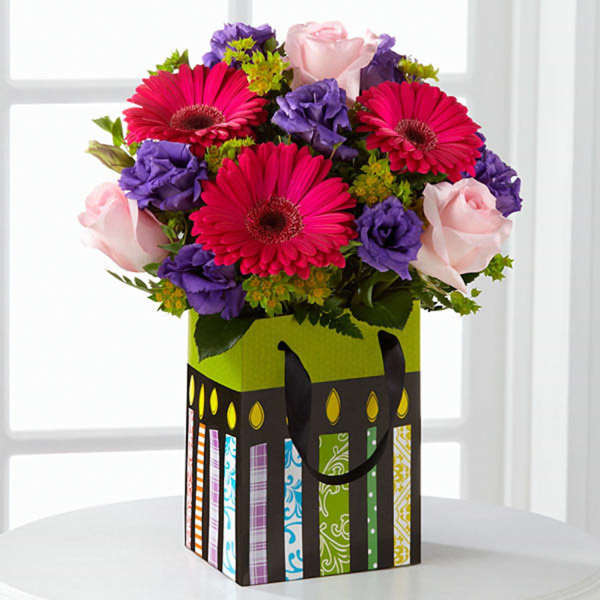Bouquet of pink gerbera daisies, purple flowers, and pale pink roses in a decorative gift bag