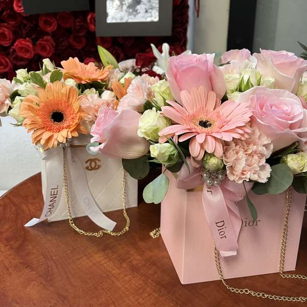 Two pastel flower arrangements in branded gift boxes with ribbon and chain handles.