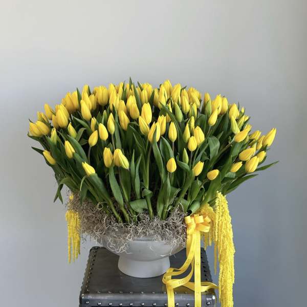 Yellow tulips arranged in a gray pedestal vase with a yellow ribbon
