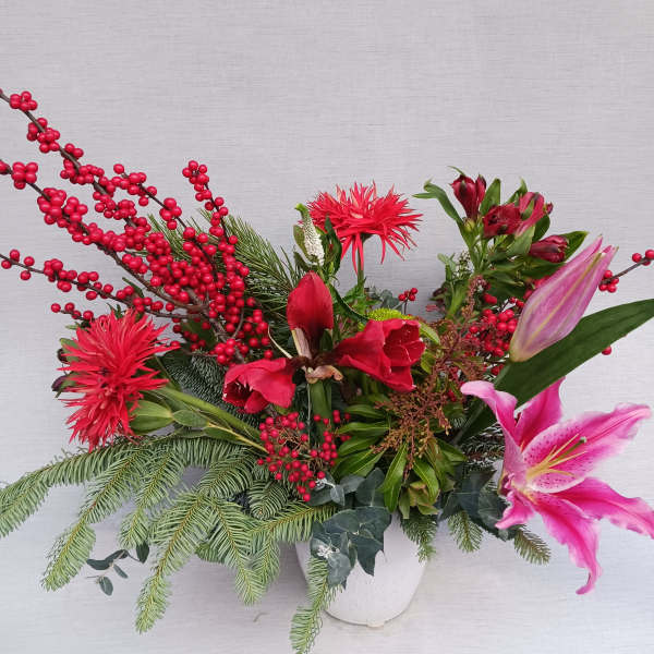 Holiday arrangement with red flowers, berries, a pink lily, and evergreen branches in a white vase