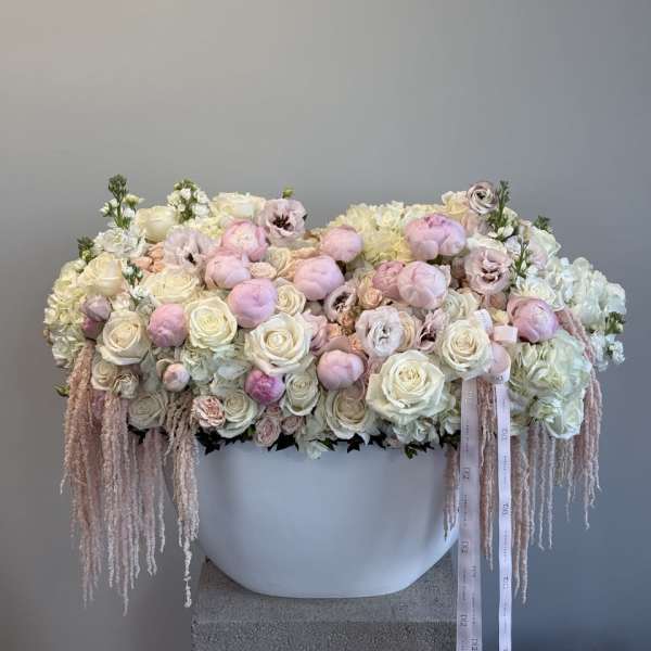 Large pastel floral arrangement in a white container with trailing ribbons