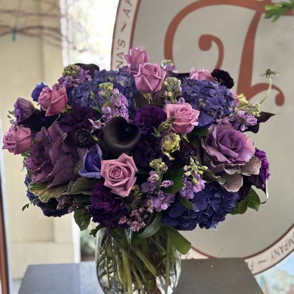 Lavender roses and purple hydrangeas arranged in a clear footed glass vase