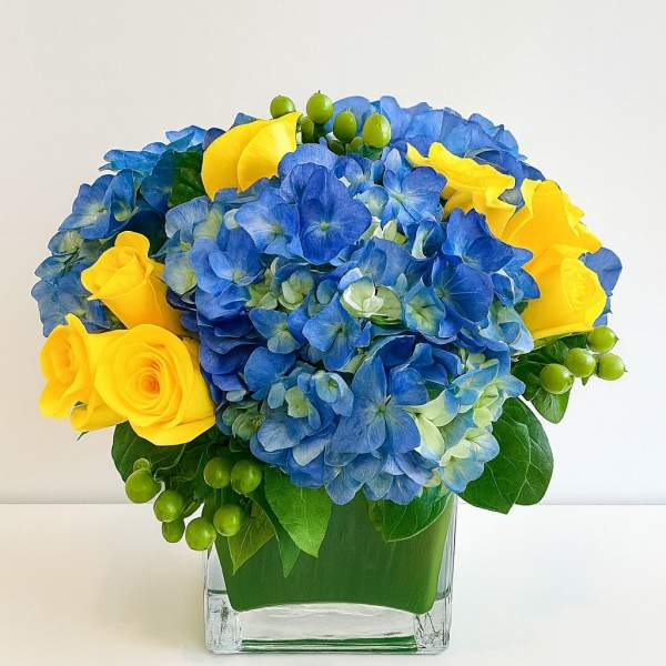 Blue hydrangeas and yellow roses in a clear square vase