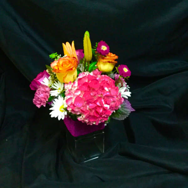 Bright mixed bouquet with pink hydrangea, orange roses, lilies, and daisies in a clear cube vase with purple wrap