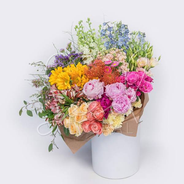 Large mixed-color bouquet with sunflowers, roses, peonies, and protea wrapped in kraft paper in a white cylinder.