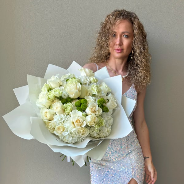 Large white and green bouquet of roses and hydrangeas wrapped in paper, held by a woman in a sequin dress