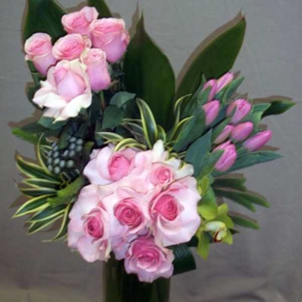 Pink roses arranged in a tall dark vase with broad green leaves.