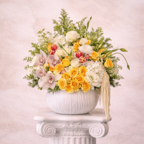 Yellow and white floral arrangement with roses, orchids, and tulips in a round white vase on a pedestal.