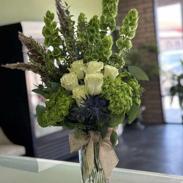 Tall green and white floral arrangement in a glass vase with a burlap bow