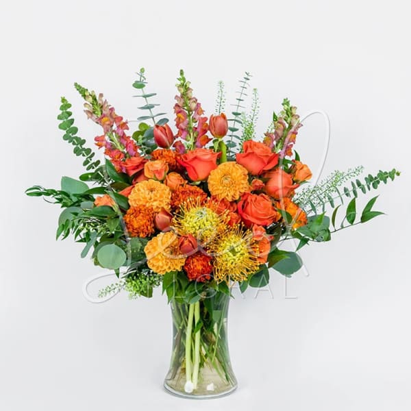Tall arrangement of orange roses, marigolds, tulips and protea in a clear glass vase