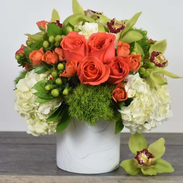 Low arrangement of orange roses, green orchids, and white hydrangeas in a white vase with one orchid bloom beside it.