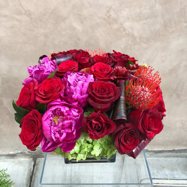 Red and pink flower arrangement with roses and peonies in a low container