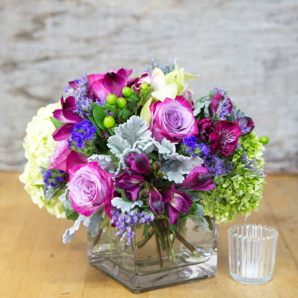 Bouquet of pink and purple flowers in a square glass vase beside a small candle