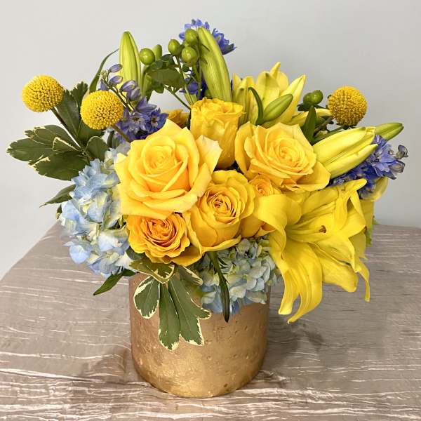 Yellow roses, lilies, and blue hydrangeas arranged in a gold vase on a light cloth-covered table